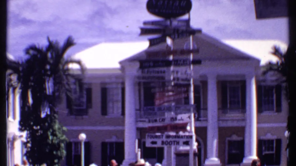 1969 Bahamas frame with island directional signage, palms, and a white-columned tourist-area building.