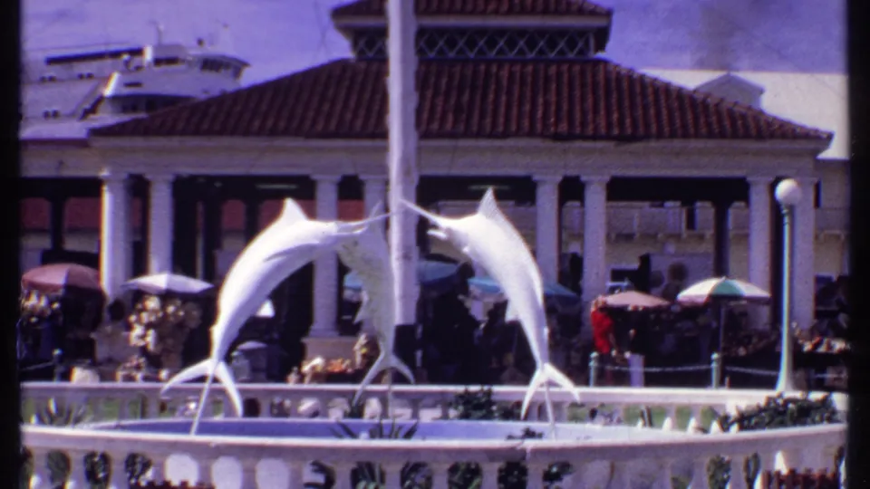 1969 Bahamas plaza view with a white marlin fountain statue, umbrellas, visitors, and a cruise ship in the background.