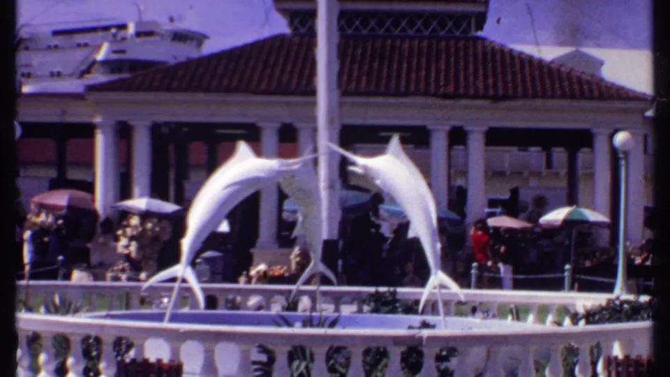 1969 Bahamas home-movie frame showing the marlin fountain statue, plaza visitors, umbrellas, and waterfront architecture.