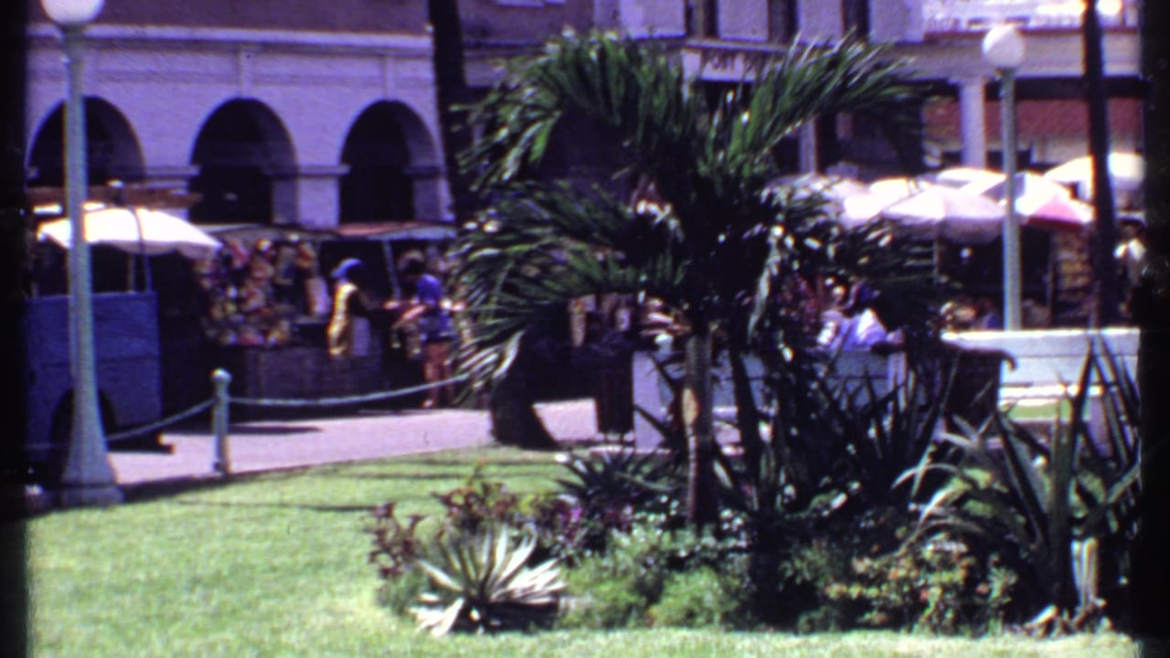 1969 Bahamas home-movie frame showing palms, a lawn, market umbrellas, pedestrians, and a colonial-style building edge.