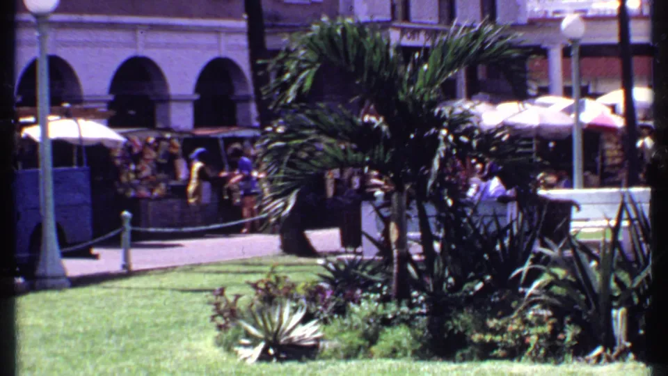 1969 Bahamas home-movie frame showing palms, a lawn, market umbrellas, pedestrians, and a colonial-style building edge.