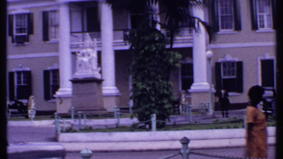 1969 Bahamas frame with a statue before a columned building, pedestrians, chain barriers, and parked cars along the street.