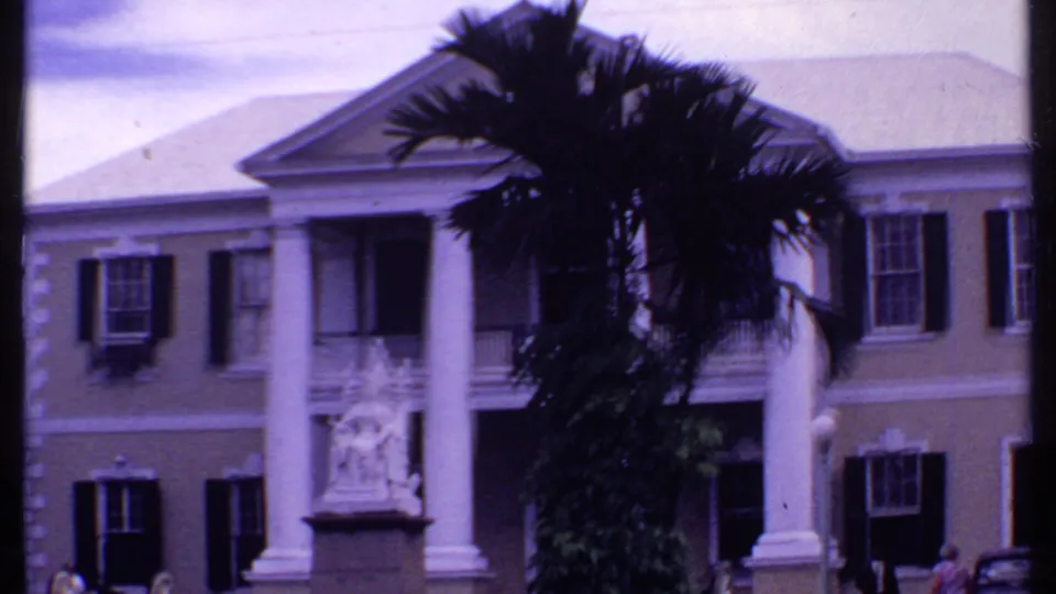 A soft 8mm view of a Bahamas colonial-style facade with white columns, a statue, palm tree, shuttered windows, and cars.