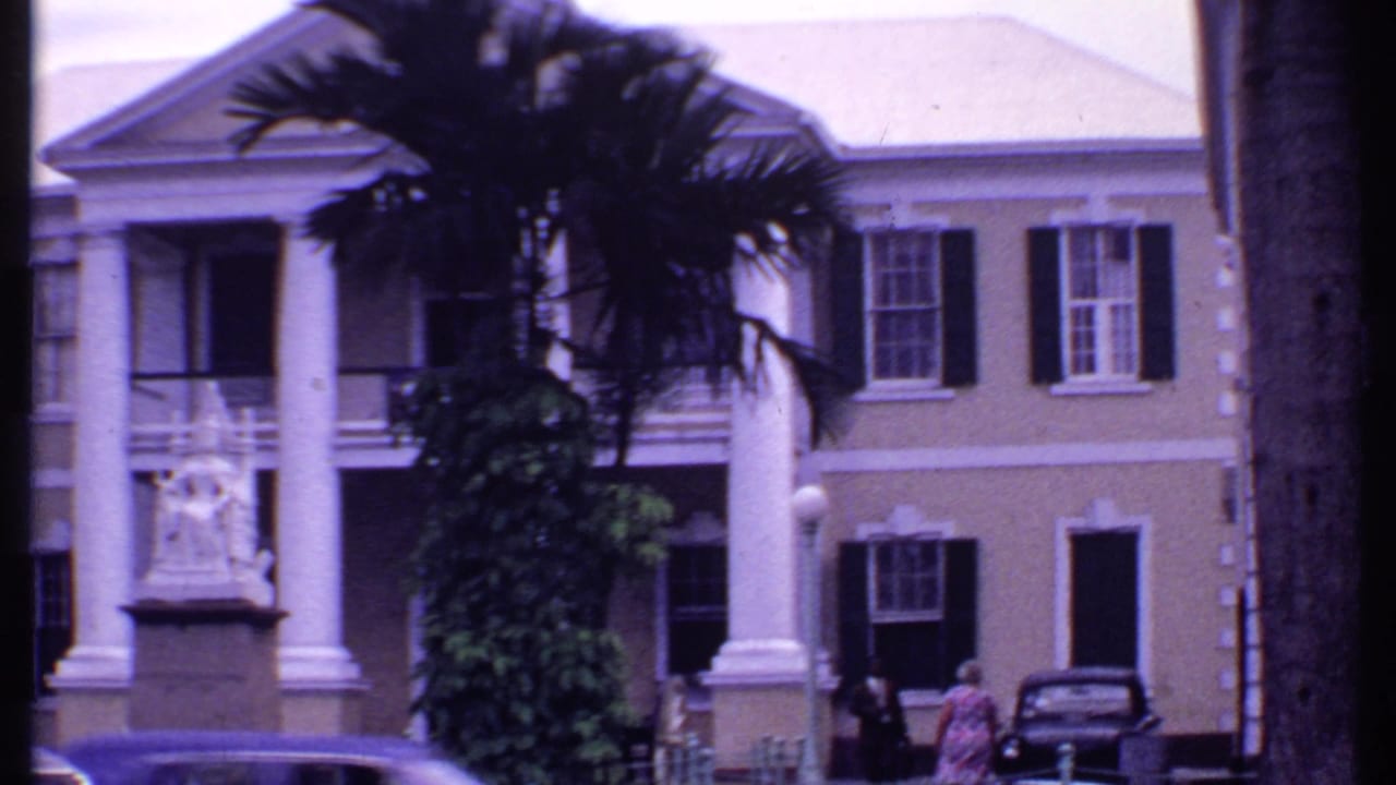Colonial-style building with white columns, palm trees, and vintage cars along a town street.