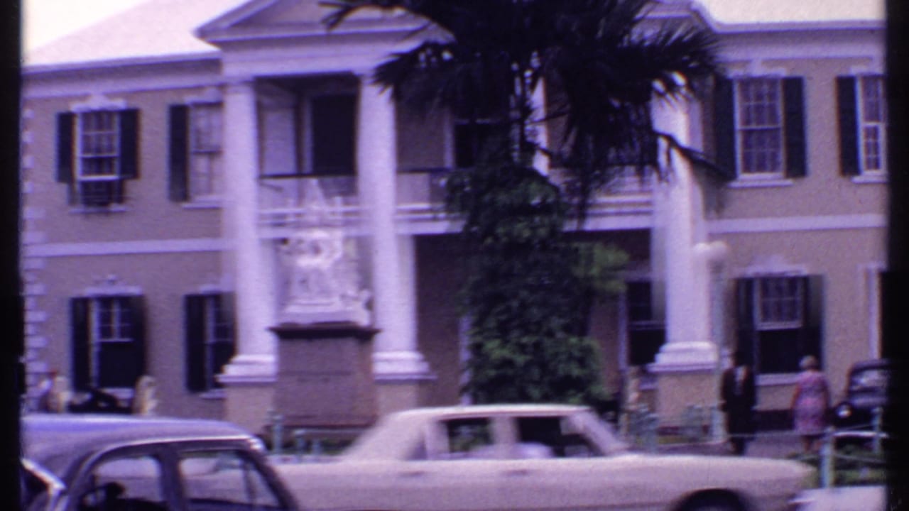 Columned building frontage with palm trees, vintage cars, and a light-colored car passing.
