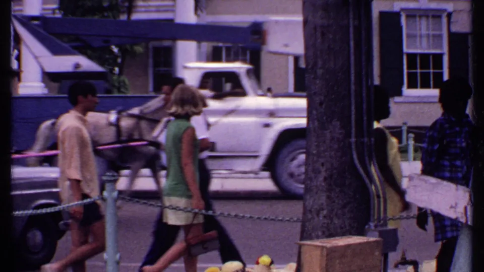 1969 Bahamas street scene with pedestrians in summer clothing, a vintage car, palms, and a columned building across the road.