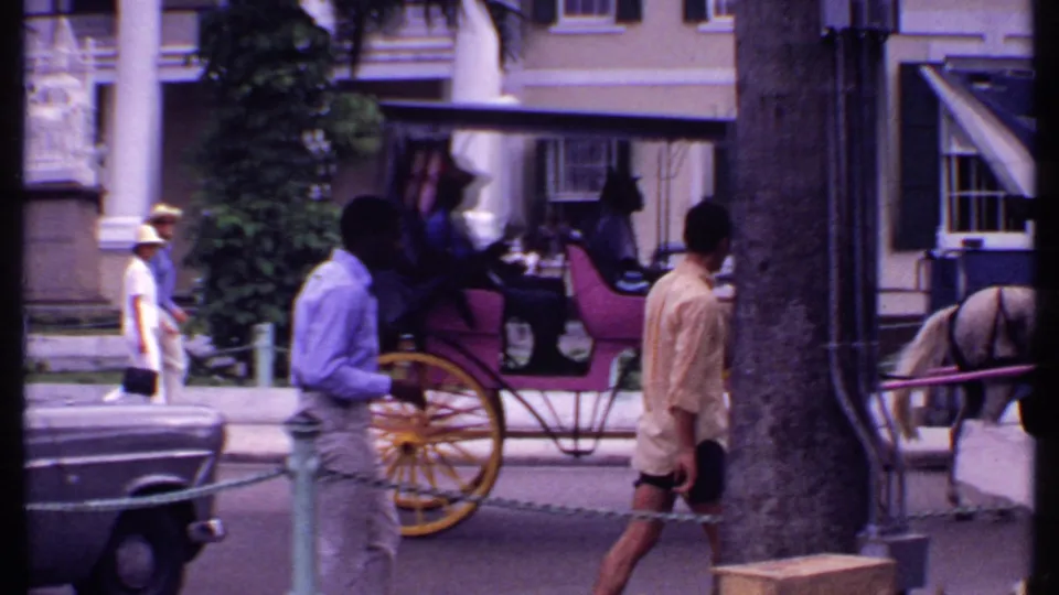 1969 Bahamas street view showing a horse-drawn carriage with passengers, pedestrians, cars, palms, and columned building details.