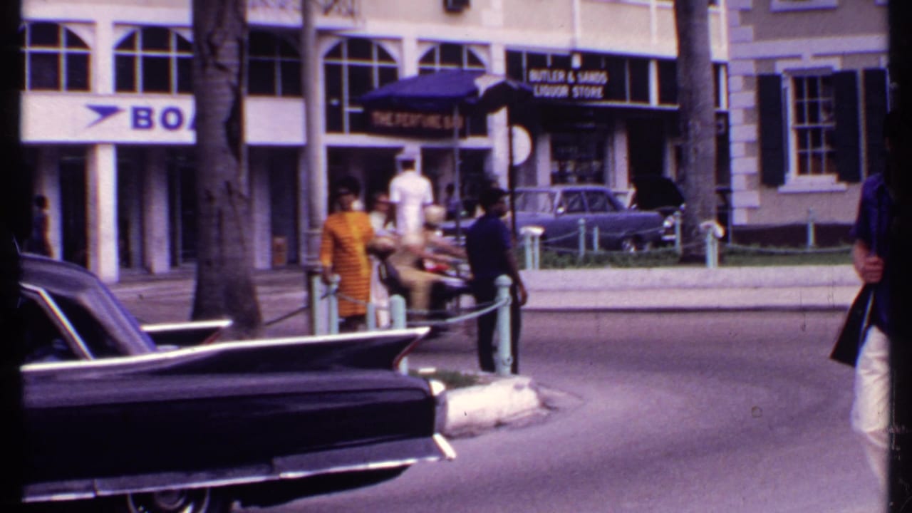 1969 Bahamas street scene with a traffic control officer, pedestrians, classic cars, palm trunks, and storefront signs.