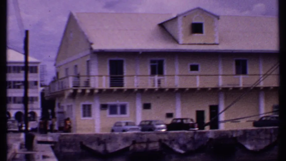 1969 Bahamas waterfront view with a pale building, balcony railings, parked cars, and the edge of a docked cruise ship.