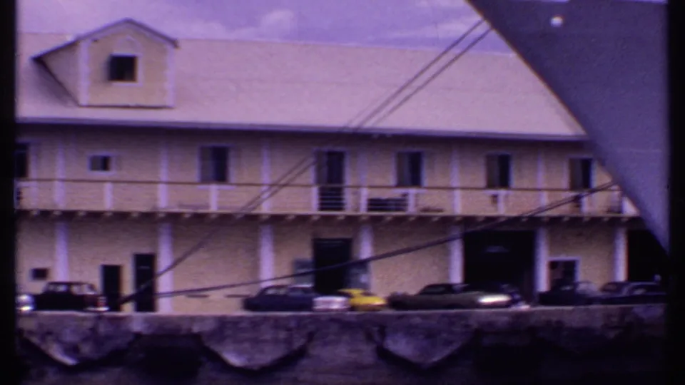 1969 Bahamas 8mm frame showing a waterfront building with upper balcony, parked vintage cars, and mooring lines crossing the view.