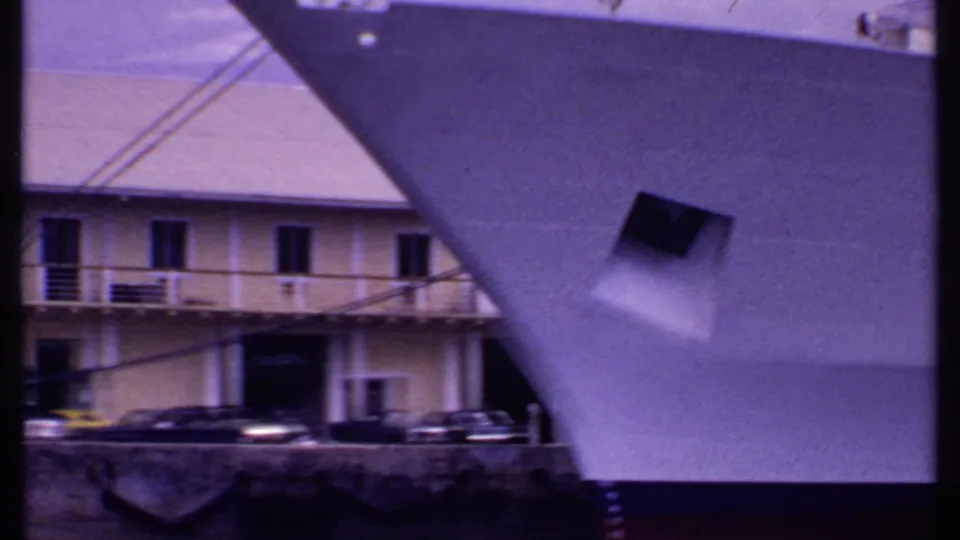 1969 Bahamas waterfront frame with the bow of a white ship crossing the foreground and buildings and cars beyond it.