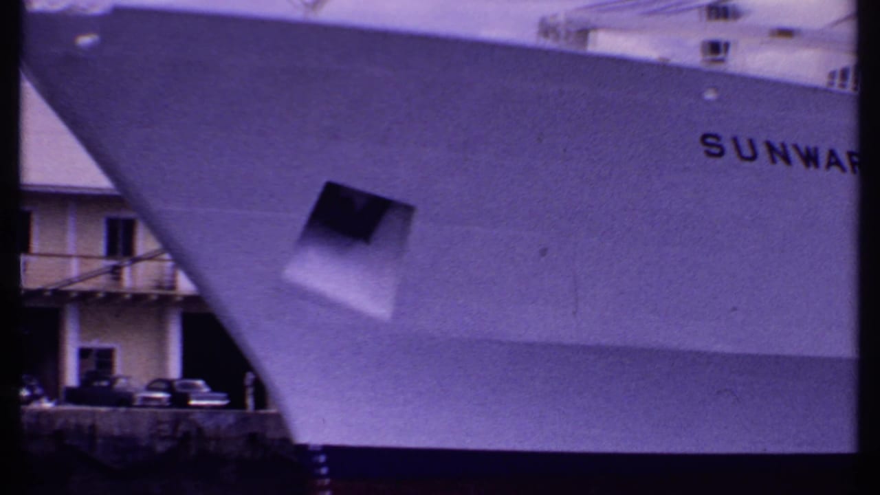 1969 Bahamas 8mm frame showing the bow of the white cruise ship Sunward at dock, with parked cars and a building behind it.
