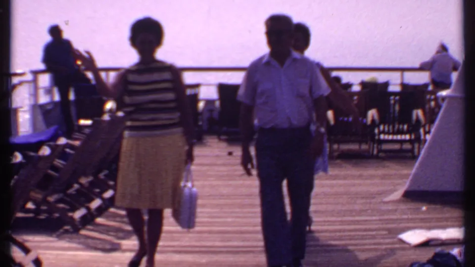 On the Bahamas ship deck, the group walks closer to the camera while the woman in a blue patterned dress raises her hand in greeting.