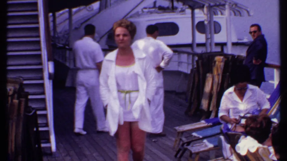 A woman in a white cover-up and shorts walks toward the camera on a 1969 Bahamas cruise ship deck with other passengers nearby.