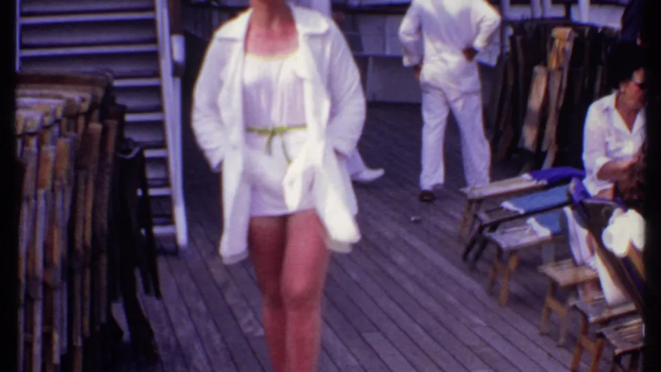 The woman in a white cover-up walks closer to camera on the cruise deck, with wooden chairs, stairs, and seated passengers visible.