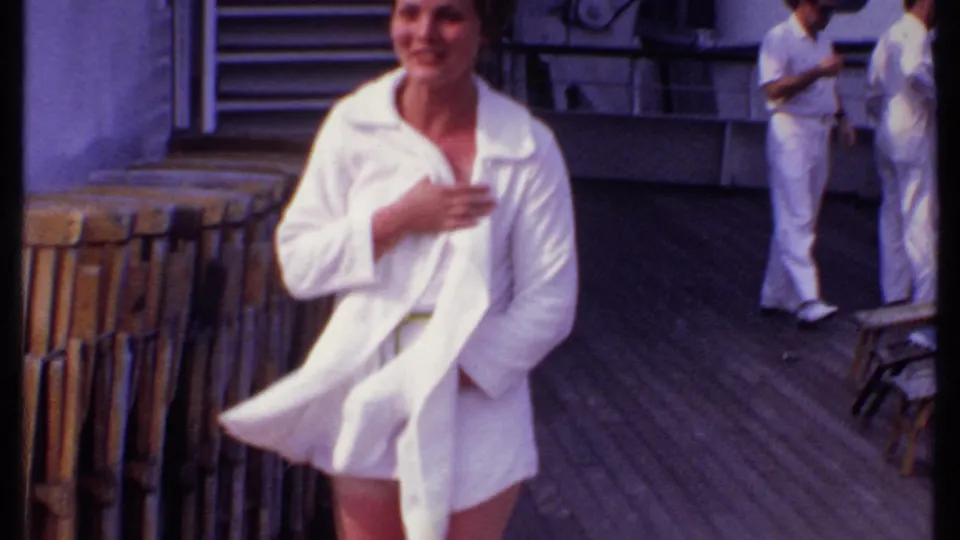 A woman in a white cover-up walks and smiles on the 1969 Bahamas cruise deck, with passengers and stacked deck chairs behind her.