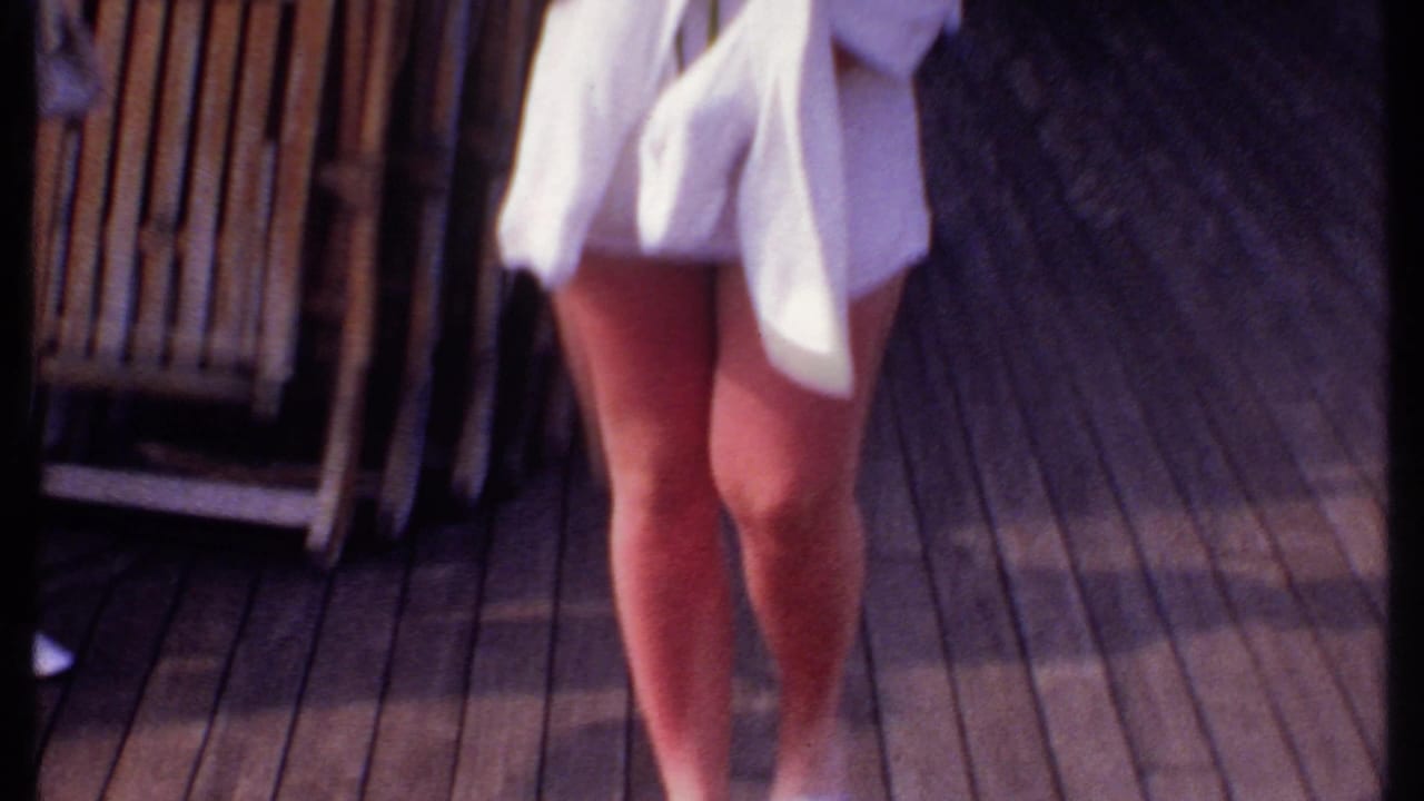 In Bahamas 1969 home-movie footage, an adult woman in white shorts crosses a wooden ship deck beside deck chairs.