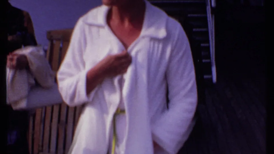 An adult woman in a white blazer-like coverup adjusts the garment while moving along a Bahamas ship deck in 1969.