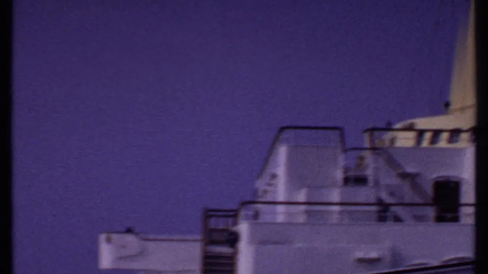 The camera turns to the ship's white upper structure and railings, rendered in grainy 8mm color against a purple sky.