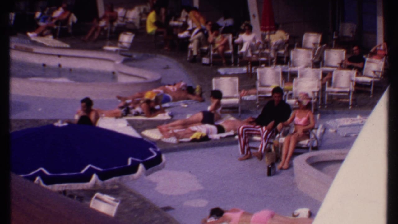 Guests sit and recline on lounge chairs beside a curved resort pool under a large blue umbrella.