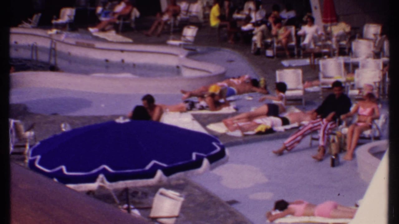Several people relax by the pool, including a seated man in red-and-white striped pants near lounge chairs.