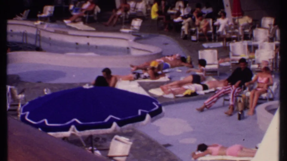Bahamas poolside home-movie frame showing seated resort guests, sunbathers, lounge chairs, and a man in red-and-white striped pants.