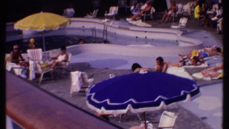1969 Bahamas resort pool area with a blue umbrella foreground, yellow umbrella near the water, lounge chairs, and guests relaxing around the deck.
