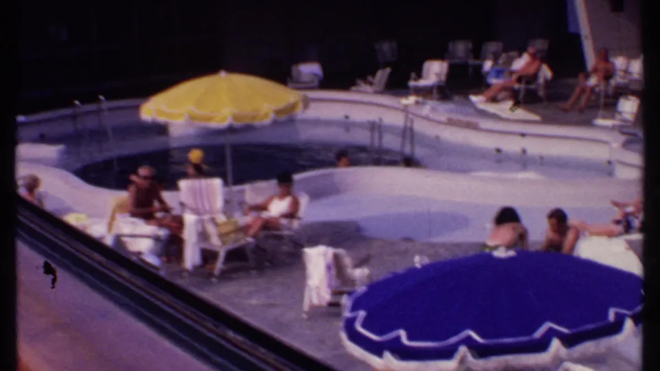 Bahamas resort poolside frame with curved pool, yellow and blue umbrellas, lounge chairs, towels, and guests sitting or reclining in swimwear.