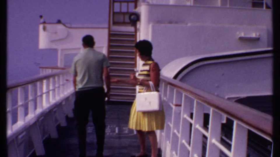 The woman appears to gesture toward the man on the Bahamas cruise deck, with white railings and stairs visible in the 8mm frame.