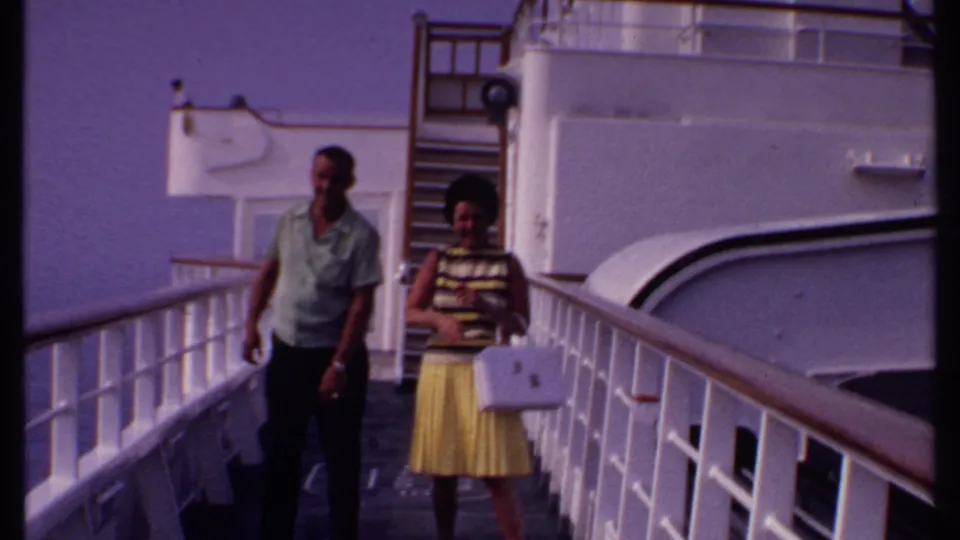 1969 Bahamas home-movie frame of the couple walking along the cruise ship deck, with the woman looking down at her purse.