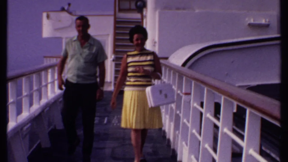 A man in a light shirt and a woman in a yellow skirt walk toward the camera on a sunlit cruise ship deck in the Bahamas footage.