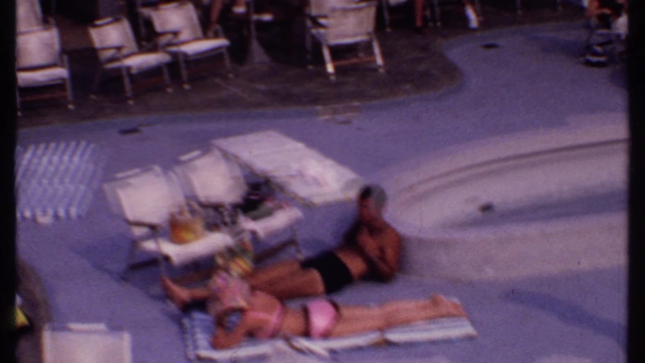 Adults sunbathe on lounge chairs and towels beside a curved resort pool.