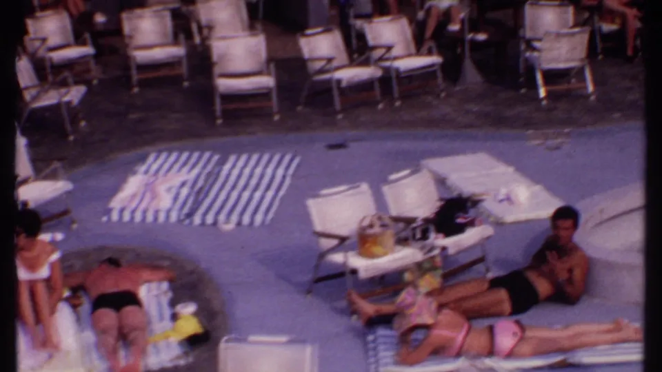 Adults at a Bahamian resort pool in 1969 rest on striped towels and lounge chairs in soft 8mm color.