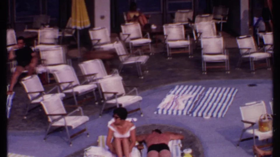 1969 Bahamas home-movie frame with a woman seated near the pool and a man lying on a striped towel.