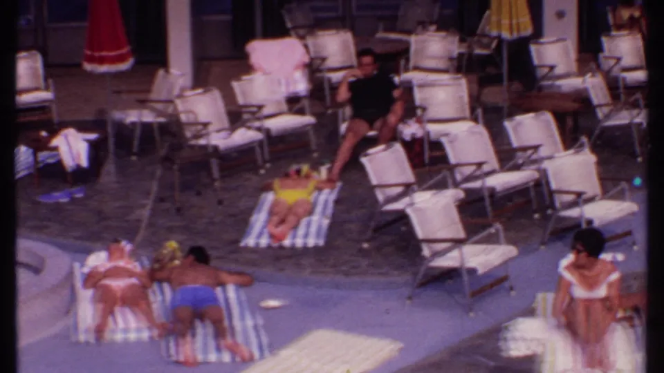 Elevated 8mm view of adults lounging beside a Bahamas resort pool in 1969, with striped towels and rows of chairs.