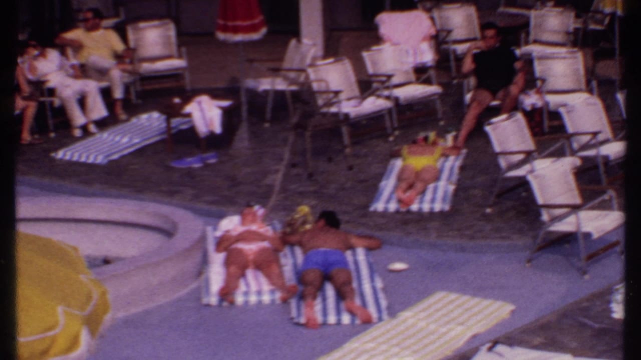 Resort pool with sunbathers on towels, lounge chairs, and a yellow umbrella in the foreground.