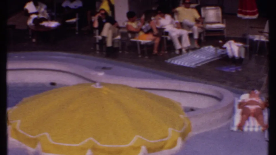 Bahamas resort pool deck in 1969 with seated guests, loungers, towels, and a large yellow umbrella partly blocking the pool.