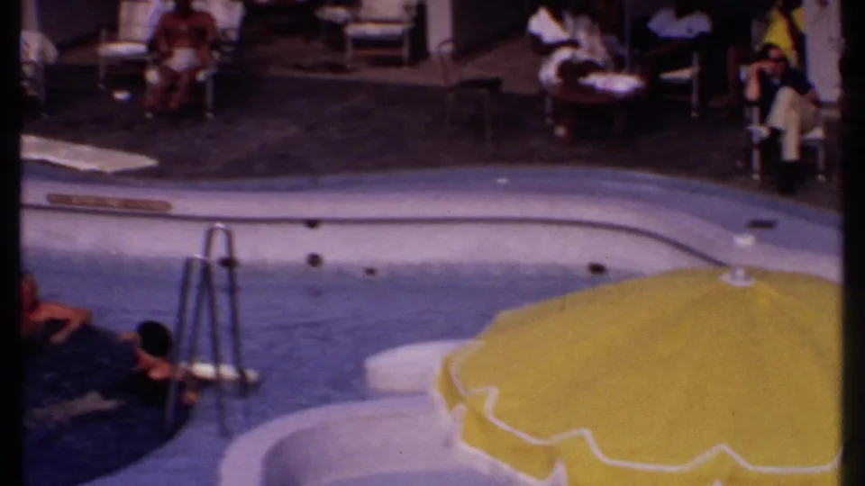 1969 Bahamas resort pool view with a metal ladder, yellow umbrella, and swimmers or guests at the edge of the water.