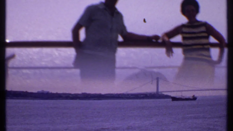 A shipboard view looks toward a distant suspension bridge and small boat on the water, partly obscured by reflection or double exposure from the deck scene.