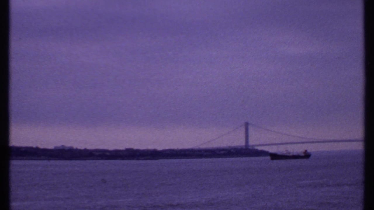 Suspension bridge across water with a dark ship in the foreground under a cloudy purple sky.