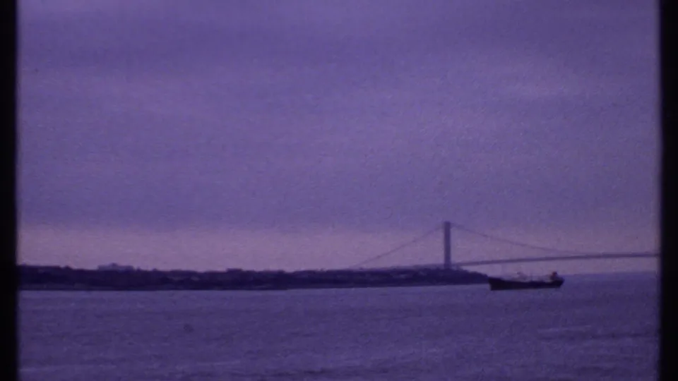 1969 8mm frame of a suspension bridge over water, with a large dark ship below and a cloudy purple-blue dawn sky.