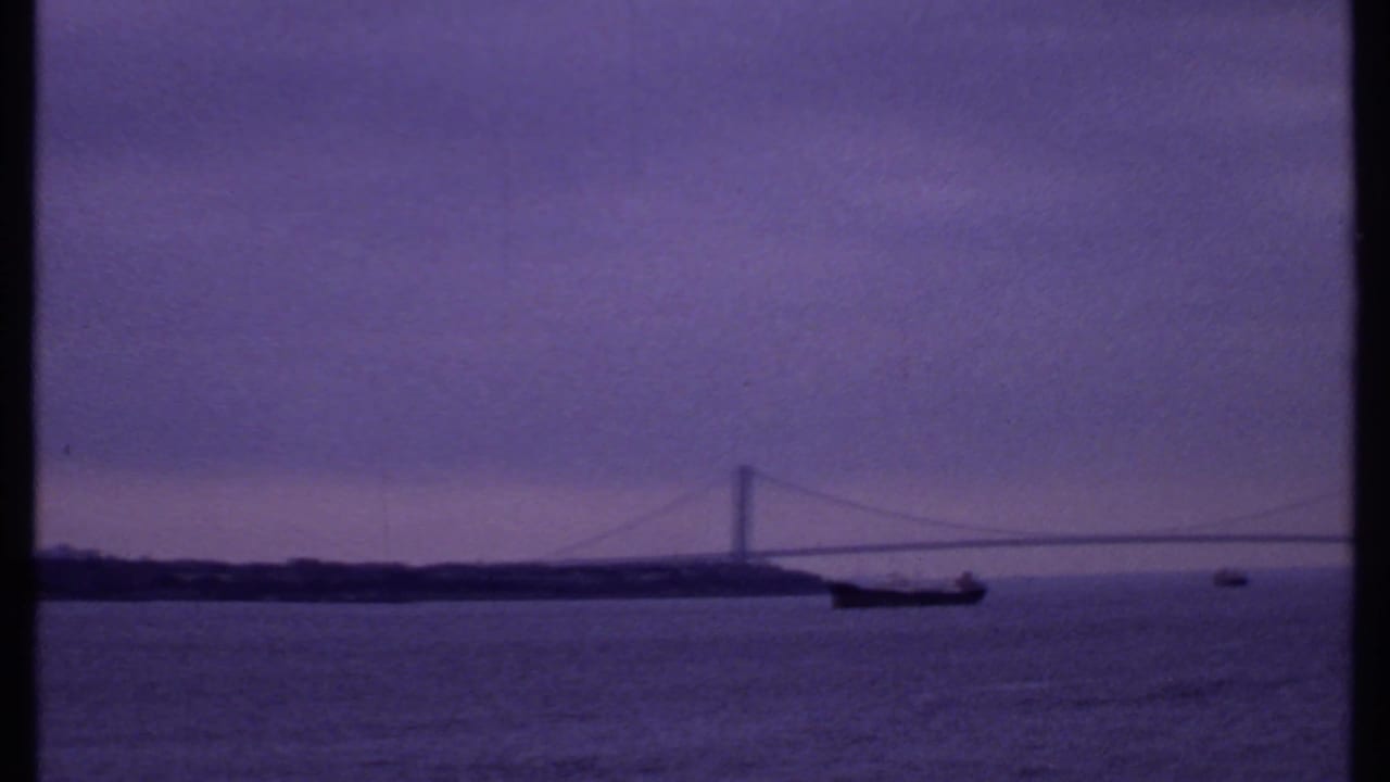 1969 8mm view of a suspension bridge over a broad waterway, with a dark ship near the span and cloudy dawn light.