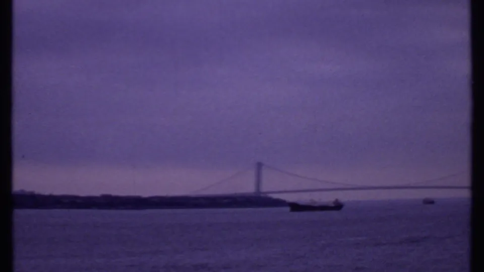 1969 8mm view of a suspension bridge over a broad waterway, with a dark ship near the span and cloudy dawn light.
