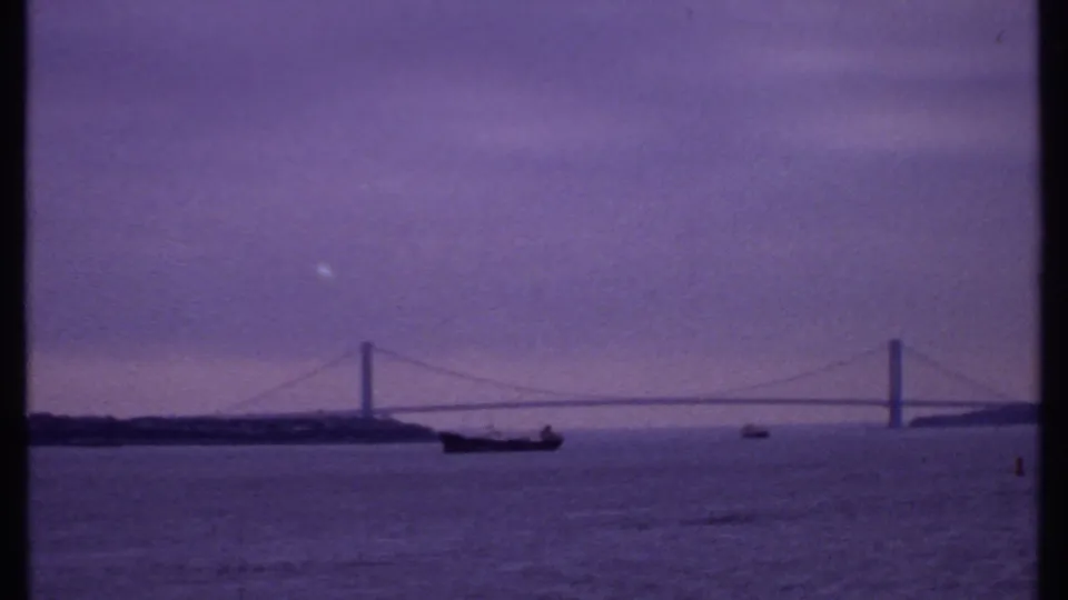 1969 8mm frame showing a wide harbor or bay view with a suspension bridge, a large dark ship, and a smaller distant boat.