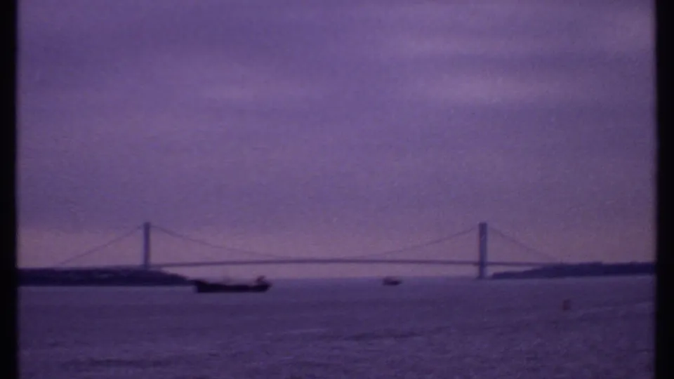 1969 8mm frame of a distant suspension bridge spanning water, with ship traffic below and a purple overcast dawn tone.
