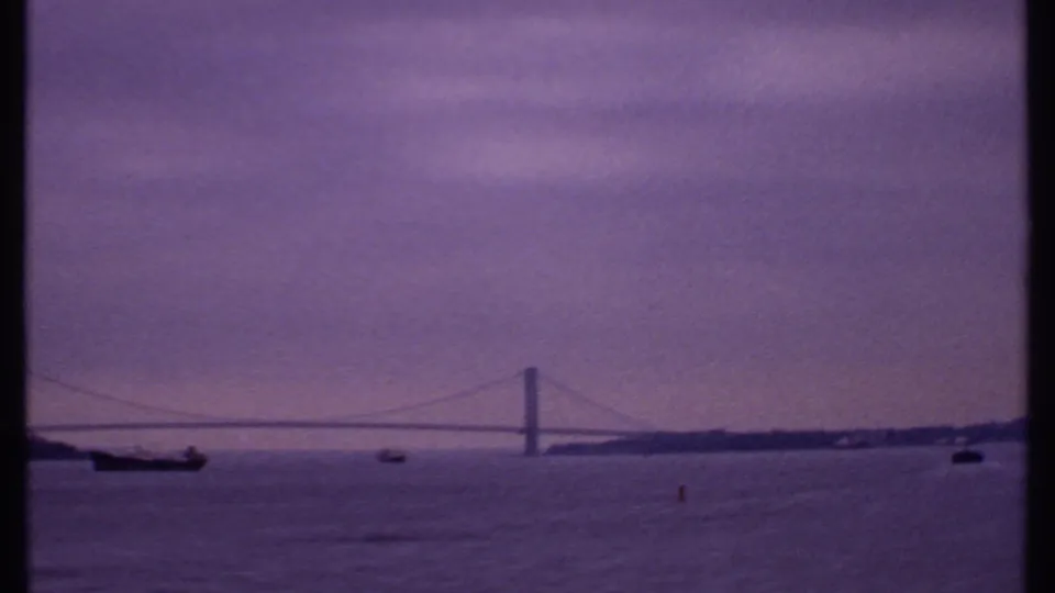 1969 Bahamas frame with bridge span, distant shoreline, cargo vessel, small boats, and soft purple evening light.