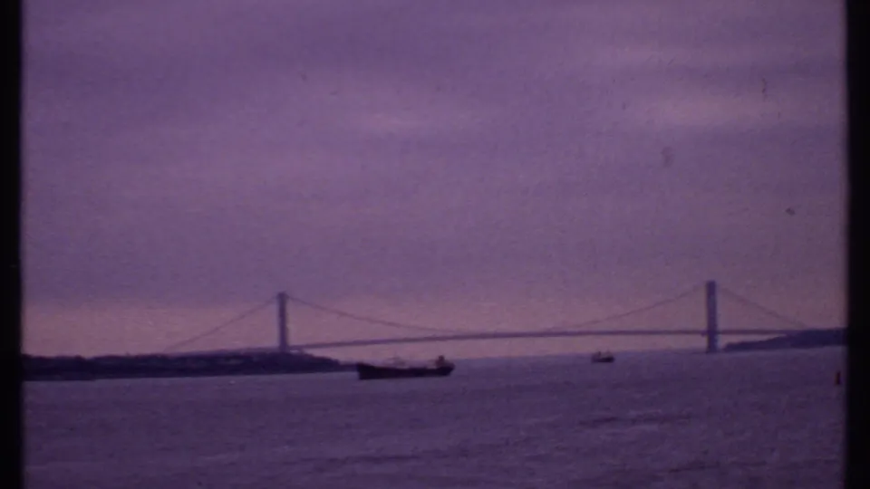 1969 Bahamas frame showing a low bridge span across open water with boats in the foreground and a purple dusk sky.
