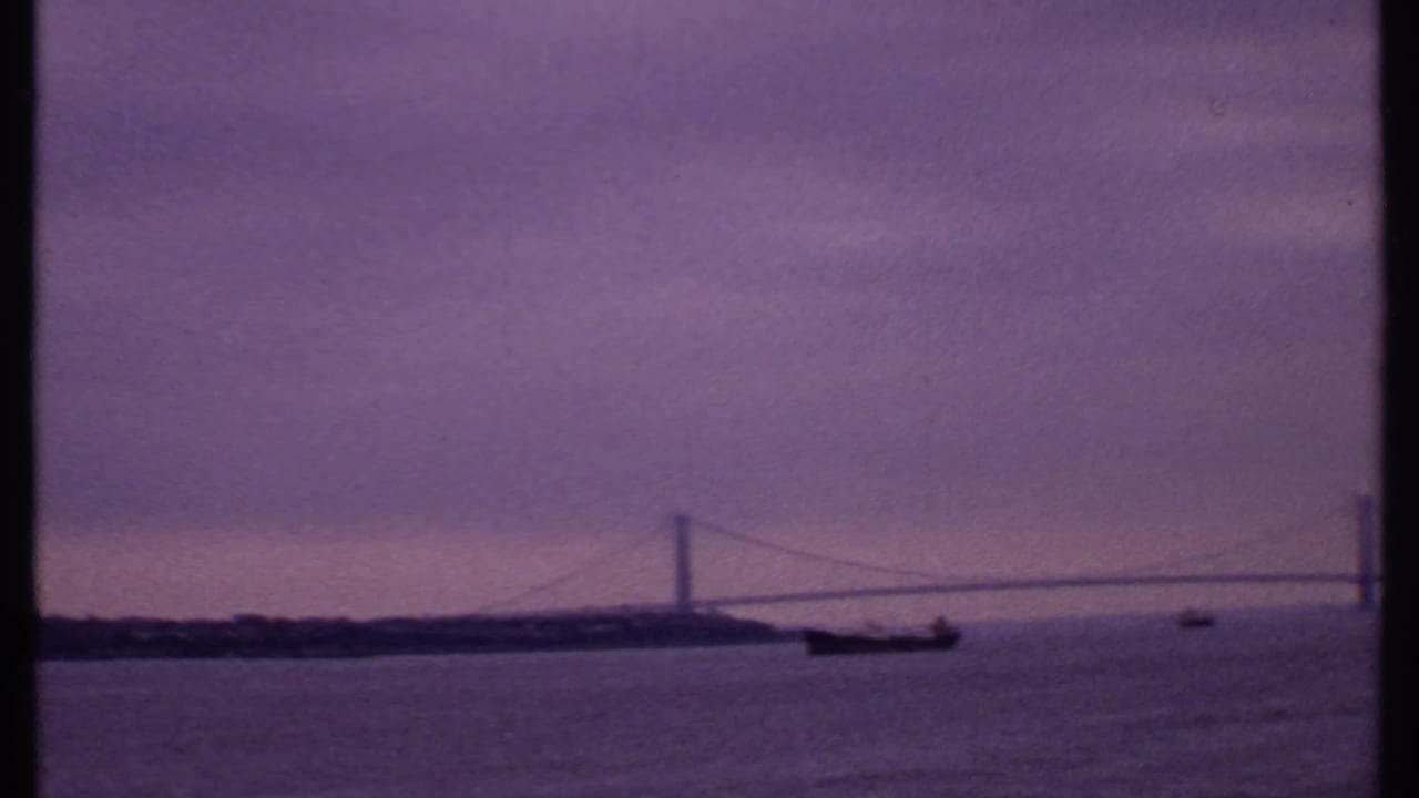 1969 8mm harbor view with a distant suspension bridge, small boats, soft skyline, and purple-tinted sky.