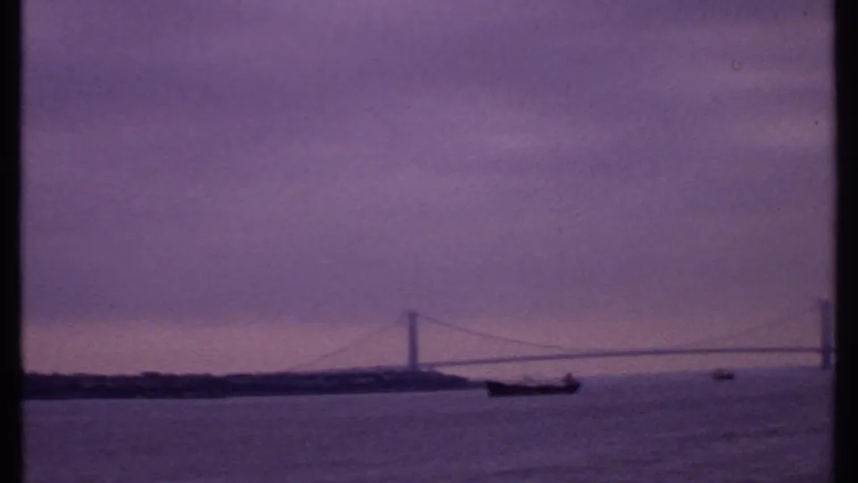 1969 8mm harbor view with a distant suspension bridge, small boats, soft skyline, and purple-tinted sky.