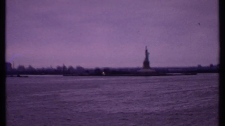 1969 8mm frame showing the Statue of Liberty at a distance, with harbor water, shoreline, and soft skyline haze.
